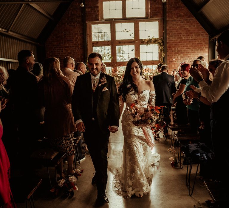 Groom in dark suit with light coloured patterned tie and pocket square and dried flower boutonniere holding hands with bride in off the shoulder bodycon wedding dress with long, sheer sleeves and lace detailing holding dried flower bouquet with roses, eucalyptus, peonies and autumn leaves walking down the aisle at The Giraffe Shed wedding venue