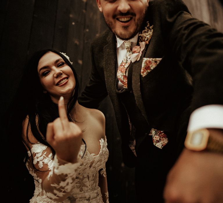 Groom in dark suit with flower patterned tie and matching pocket square with dried flower boutonniere and bride in off the shoulder bodycon wedding dress with long, sheer sleeves and lace detailing giving the middle finger