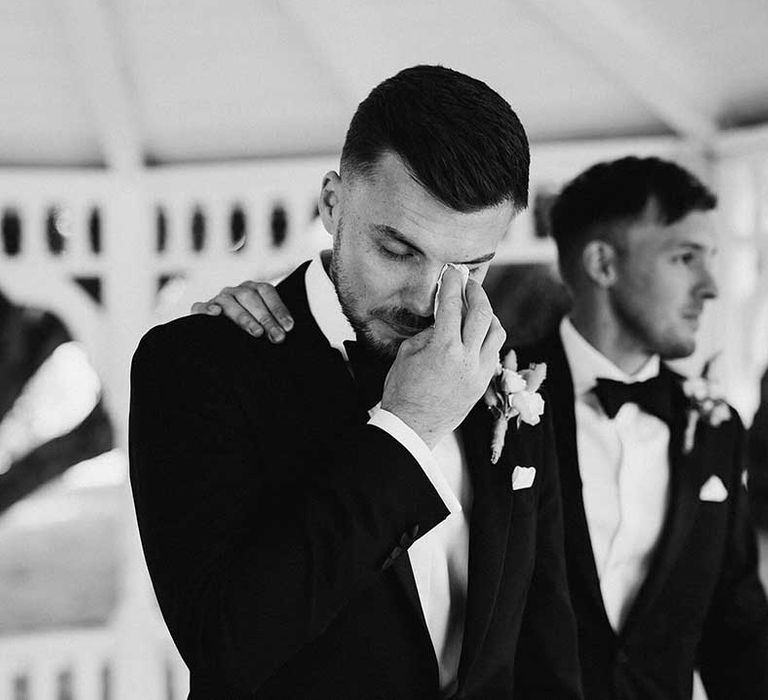 Groom in classic Ralph Lauren black tux with black bowtie and blush rose boutonniere wiping a tear at wedding ceremony 