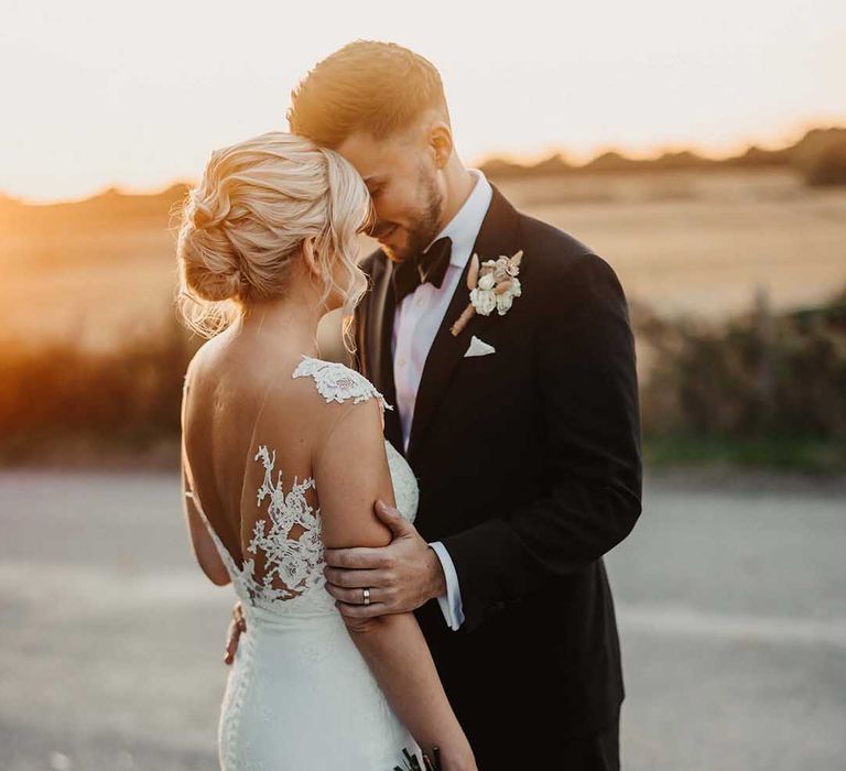 Bride in lace wedding dress with sheer, lace top and clusters of lace flowers on the shoulders with button details holding neutral blush bouquet with pink garden roses, peonies, dahlias, dried flower, Persian buttercups and foliage embracing groom in classic Ralph Lauren black tuxedo with black bowtie and dried flower boutonniere