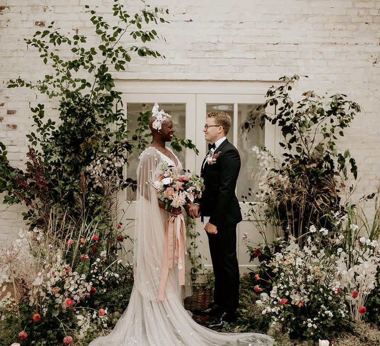 Black bride with alopecia smiling with groom in black tie with glasses standing at the altar decorated with wildflower columns 