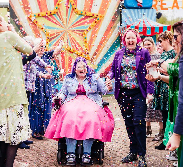 Bride in purple velvet blazer and black suit trousers with purple bowtie, pink patterned pocket square and colourful patterned shirtband bride in wheelchair wearing personalised denim jacket, pink wedding dress and blue tights doing confetti walk at theme park wedding 