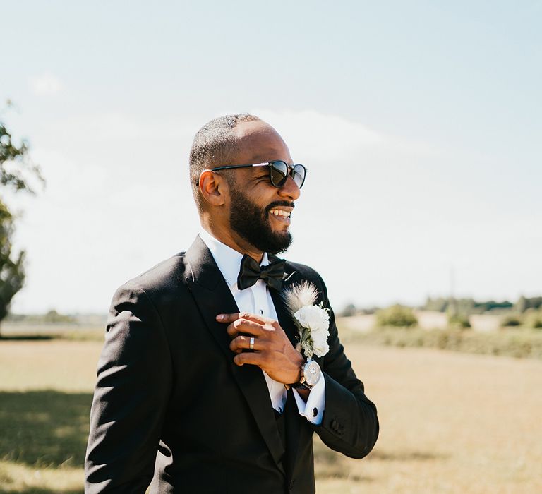 Groom wears three piece black-tie suit complete with white floral buttonhole and sunglasses 