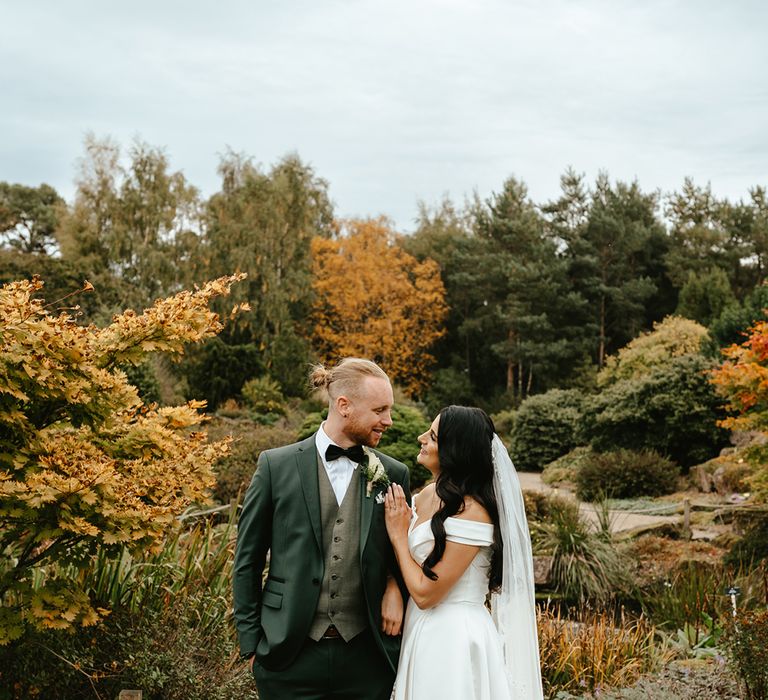 Bride in off-the-shoulder wedding dress looks lovingly toward her groom in green suit during outdoors couples portraits 