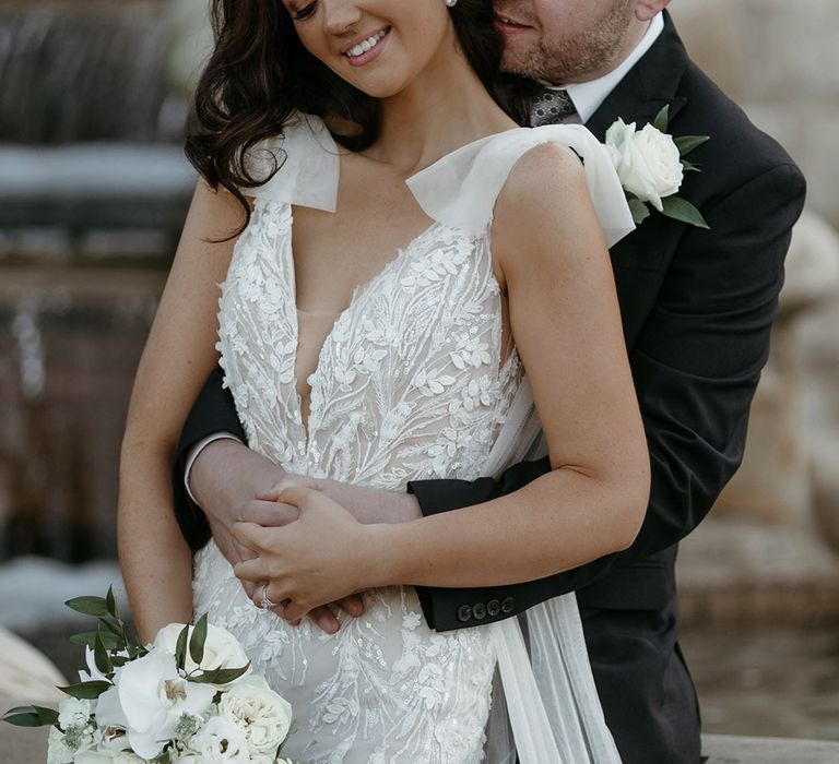 Groom in black suit with white rose buttonhole embracing the bride in a flower embellished wedding dress with cascading bouquet and tulle straps 