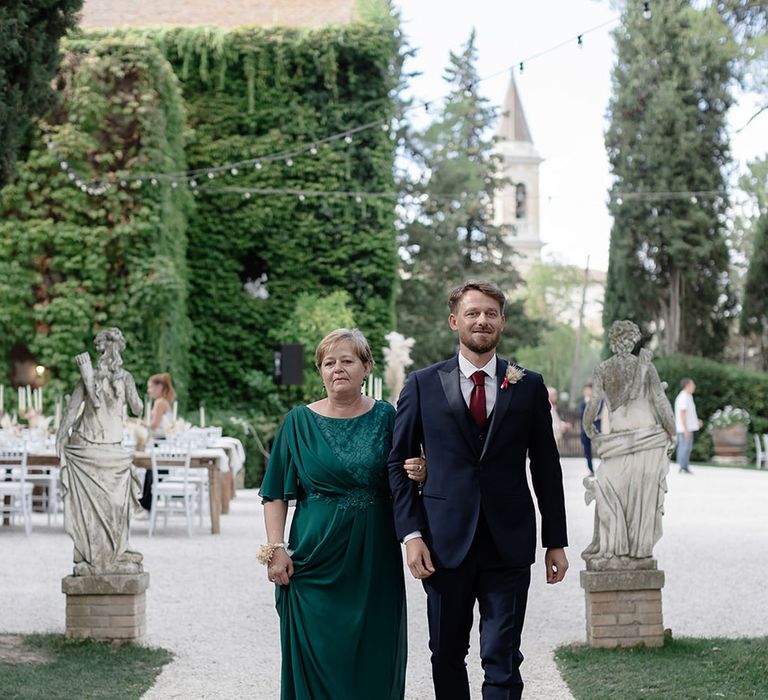 Groom in three piece suit walks alongside his mother in deep green dress 