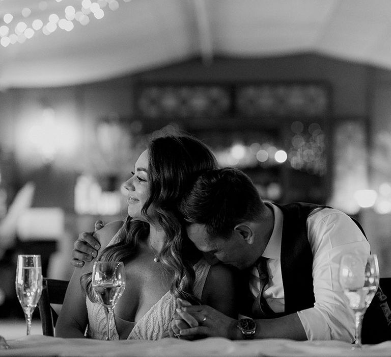 The groom kisses the bride on her shoulder as they sit at their wedding reception as they listen to speeches 