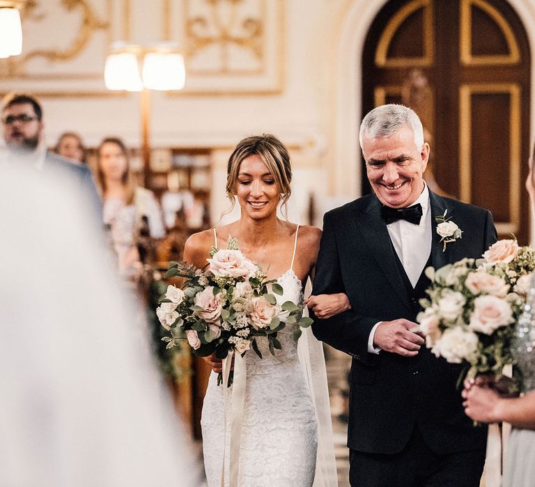 Father of the bride in traditional black tie with white rose buttonhole walking the bride in a fitted lace wedding dress down the aisle 