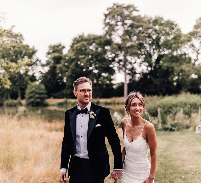 The bride wears a spaghetti strap fitted lace wedding dress smiling and strolling with the groom in black tuxedo 