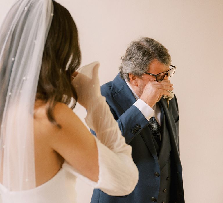 Father of the bride in a three piece navy suit wiping his eyes as he cries seeing the bride in a pearl embellished bridal outfit 