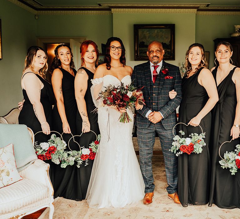 Father of the bride in a navy and orange checkered suit with the bride in an off the shoulder lace wedding dress and the bridesmaids in black satin dresses with heart shaped bouquets