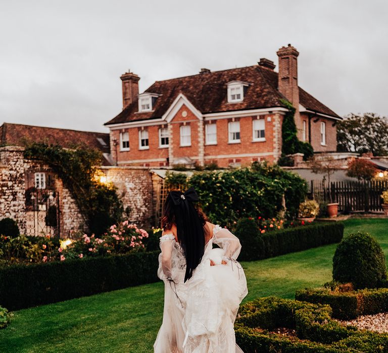 The bride in an off the shoulder embellished lace wedding dress runs toward Parley Manor country house wedding venue in Dorset 