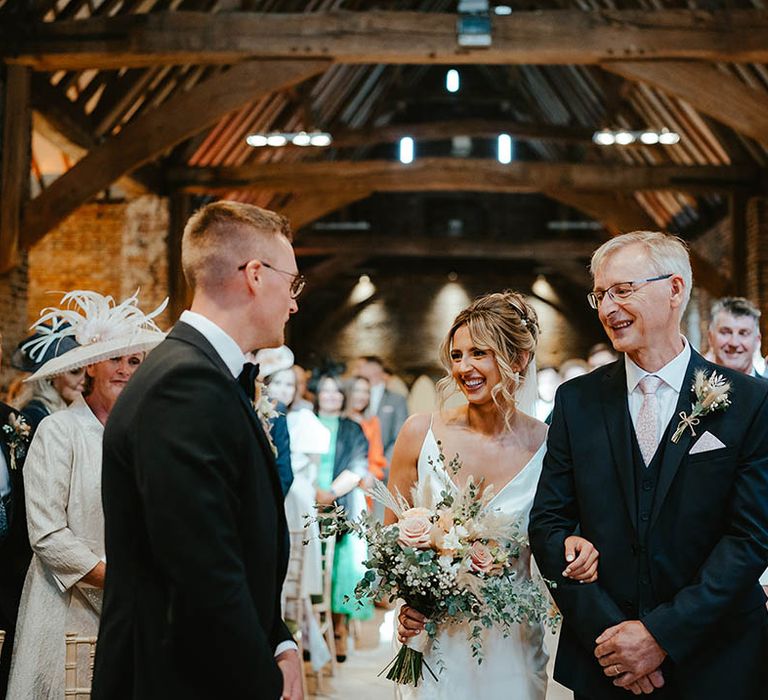 Father of the bride in three piece dark suit walks the bride in a satin wedding dress to the groom in a black tuxedo waiting at the end of the aisle 