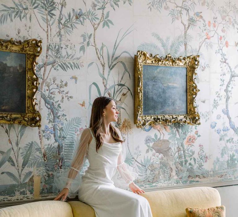 Bride in long sheer puff sleeve satin wedding dress with cowl neck sitting on yellow velvet sofa at St Giles House