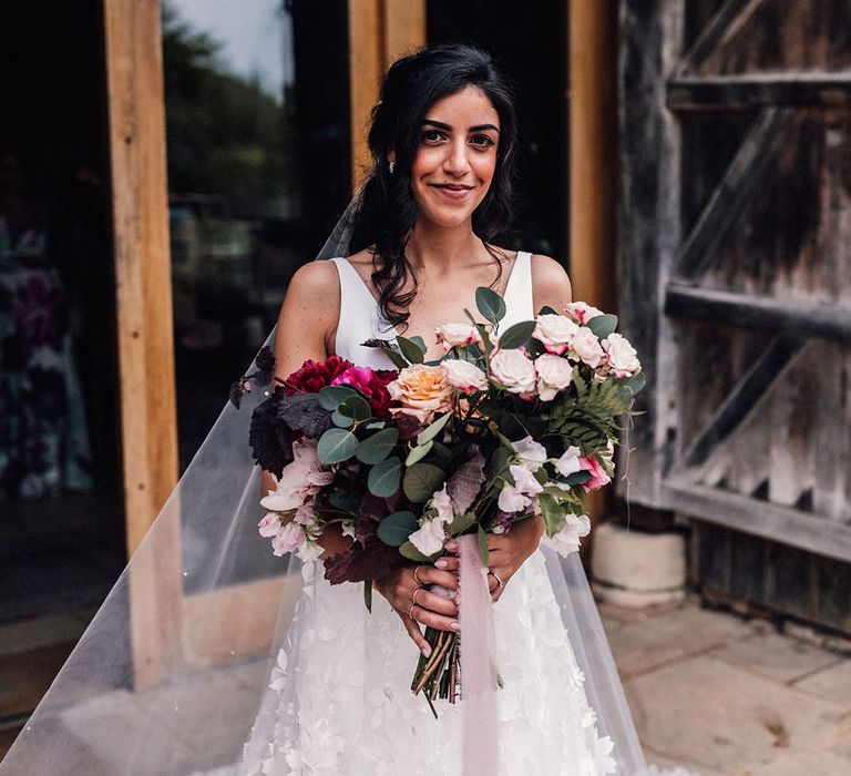 Bride holding a dark red and light pink peony and rose wedding bouquet with foliage for luxe wedding at Cripps Barn