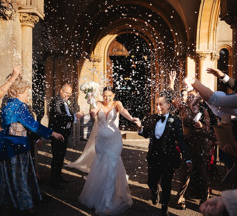 Bride in black snakeskin print tuxedo with black bowtie and white rose boutonniere holding hands with bride in lace sleeveless v neck Enzoani wedding dress with tulle finish and church length veil holding white garden rose bridal bouquet doing white petal confetti exit at wedding