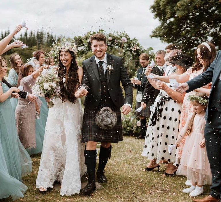 Bride in celestial 3d applique sleeveless wedding dress and flower crown doing confetti exit with groom in dark grey blazer and waistcoat, dark tartan tie with white flower boutonniere, dark grey and blue tartan kilt and wedding sporran 