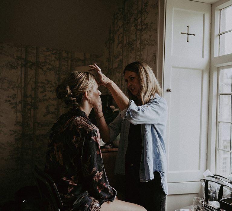 The bride sits facing a window for natural lighting as she gets her makeup done for the wedding day 