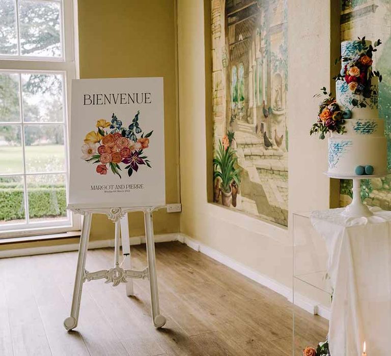 Large botanical luxe white wedding sign in French on white easel next to four tiered blue and white wedding cake 