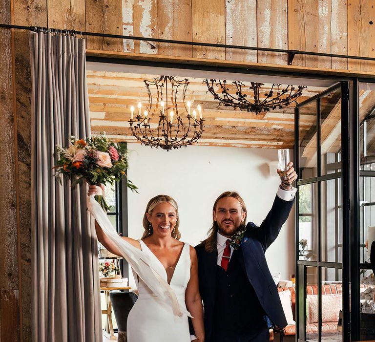 The bride in a fitted Savin London wedding dress enters the wedding reception with the groom in a three piece blue suit 