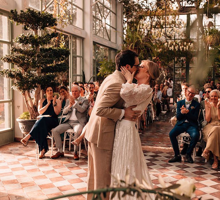 Glass house wedding venue with the bride and groom sharing their first kiss as a married couple 