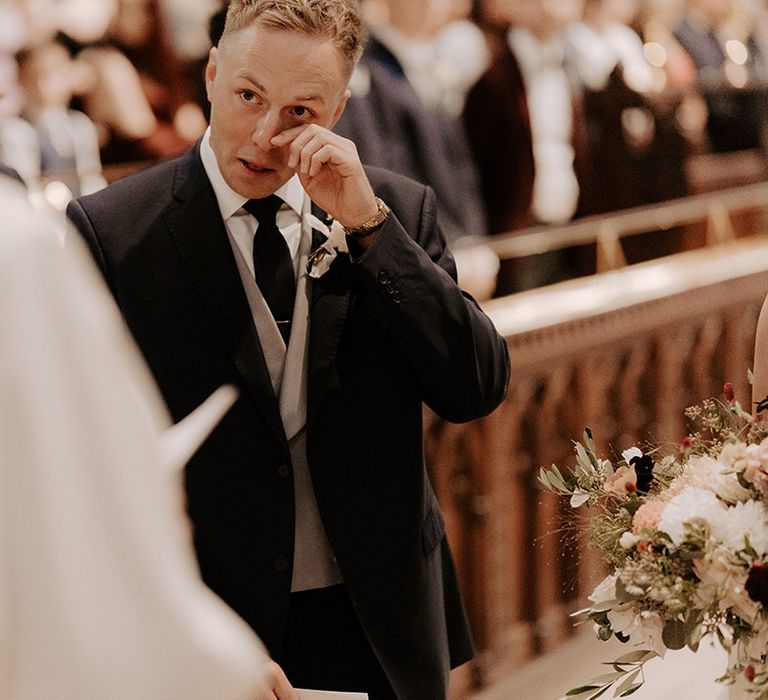 Groom in three piece suit wipes his tears as he sees the bride for the first time 