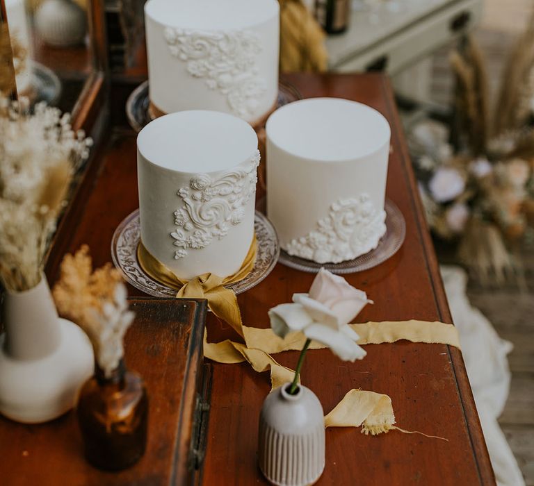 three single-tier wedding cakes with intricate design on a vintage dresser