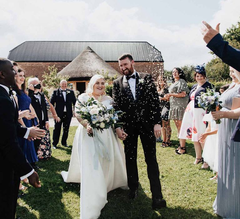 Groom in classic black grooms three piece tuxedo with white rose boutonniere and black bowtie doing colourful wedding confetti exit with bride in satin off the shoulder wedding dress holding eucalyptus, foliage, garden rose, gardenia, bluebell and dried flower bridal bouquet tied with dusky blue ribbon at Brickhouse Vineyard Devon wedding venue