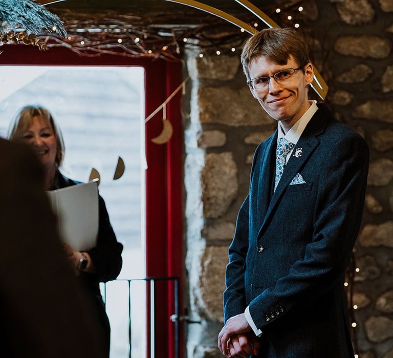 Smiling groom wearing glasses in a navy suit and floral patterned tie turns to see the bride for the first time 