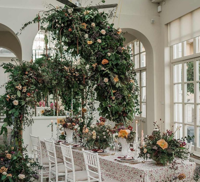 Botanical wedding tablescape with patterned floral tablecloth, twisted tapered candles, art deco vases with dried wildflower wedding centrepieces surrounded by suspended wildflower and foliage arrangements in large gold vases at Buxted Park