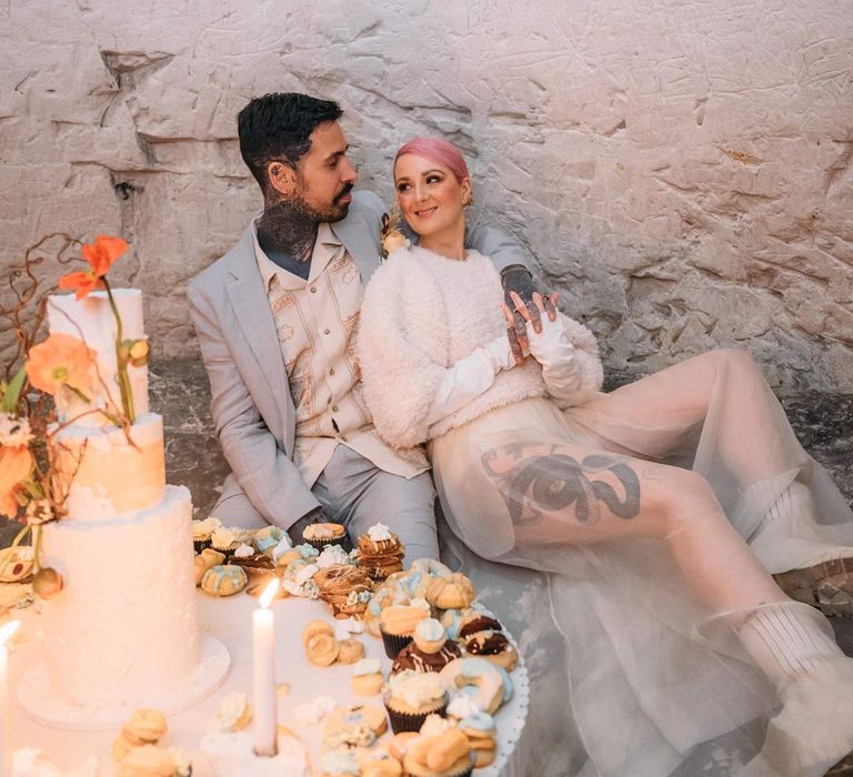 Groom in baby blue linen suit, personalised embroidered linen shirt and garden rose and pampas grass boutonniere sitting with bride in sheer overlay wedding dress and fluffy bridal coverup by wedding grazing table at Margate Caves