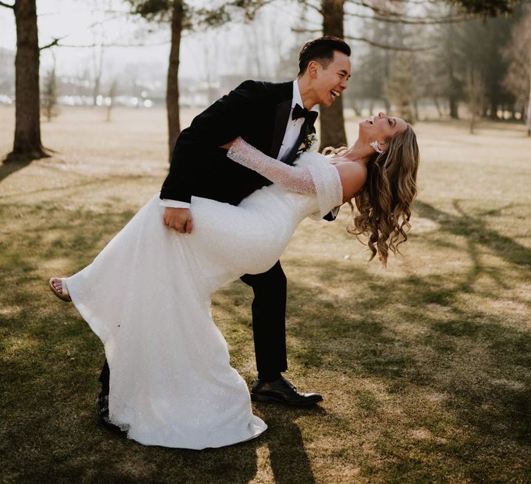 Groom in classic black tuxedo, black bowtie and white garden rose boutonniere dipping bride in off shoulder sparkly mermaid wedding dress laughing at ski wedding in France