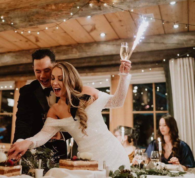 Groom in classic black tuxedo, black bowtie and white garden rose boutonniere and bride in off shoulder sparkly mermaid wedding dress cutting their Victoria Sponge wedding cake