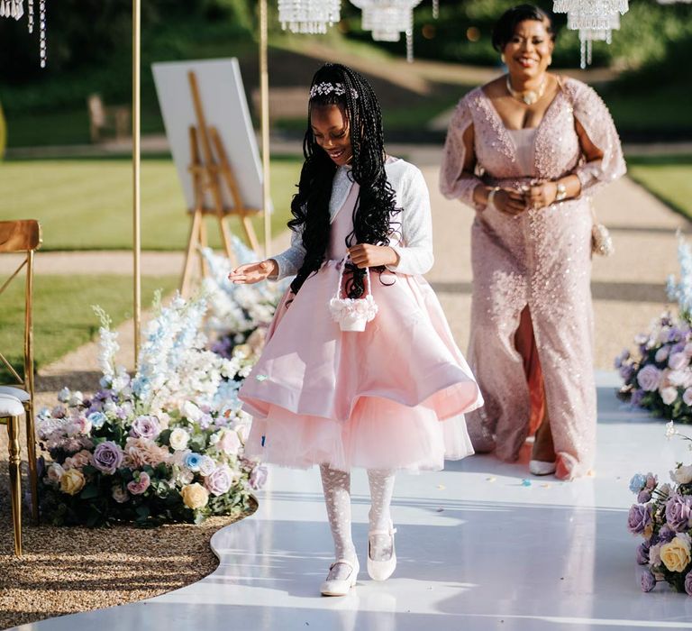 Flower girl in baby pink flower girl dress and white cardigan walking down the aisle at Syon Park wedding venue with outdoor gold and crystal chandeliers and pastel aisle flower arrangements with bride wearing illusion v-neck sheer off shoulder long sleeve pearl embellished wedding gown walking behind her