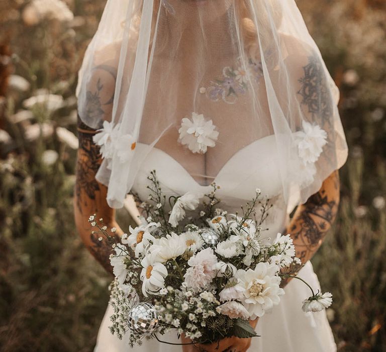 Bride with floral face paint wearing veil over her face holding white wildflower bouquet 