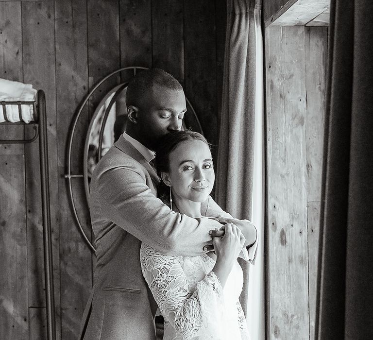 Groom embraces the bride in a long sleeve lace wedding dress as they gaze out of the window 