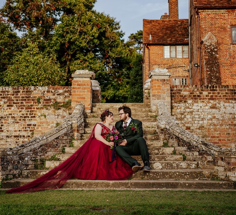 Ufton Court wedding venue with the bride and from posing on the steps for their couple portrait with the bride wearing a red wedding dress with cape