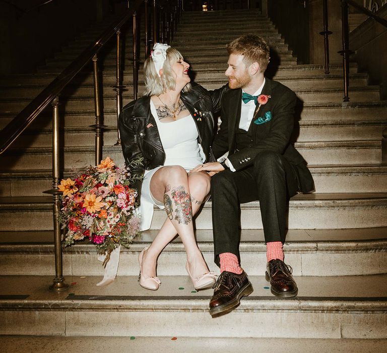 Bride in black leather jacket and short wedding dress with groom on the steps of their wedding venue 