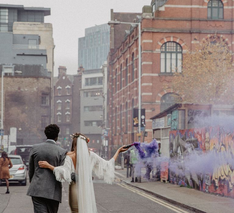 Wedding couple walk away down city street, bride holds purple smoke bomb