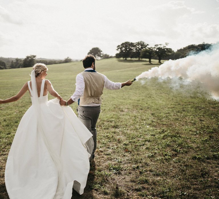 Bride in a Jesus Peiro wedding dress with back strap detail and groom in tails holding white smoke bombs in a field 