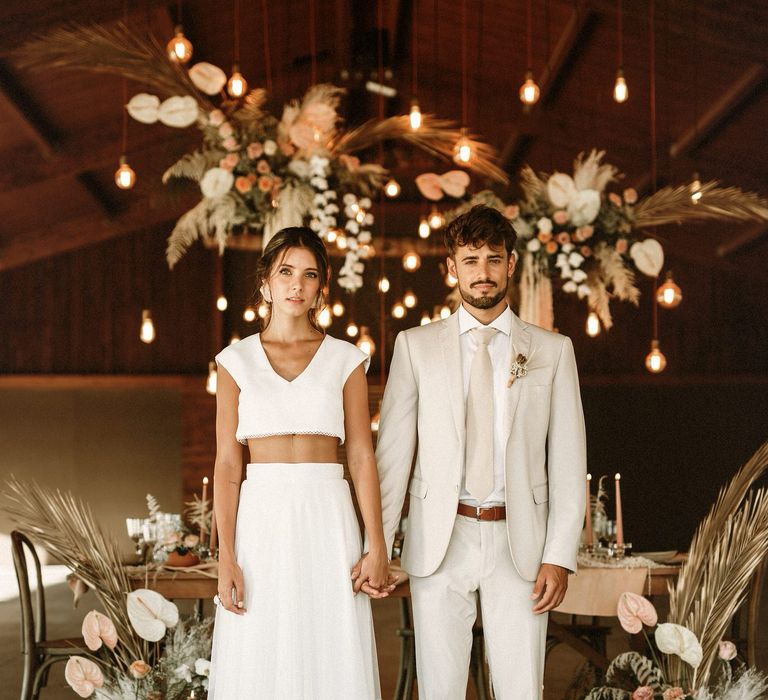 Bride in separates and groom in a beige suit standing in their intimate barn reception full of festoon lights and boho wedding flowers 