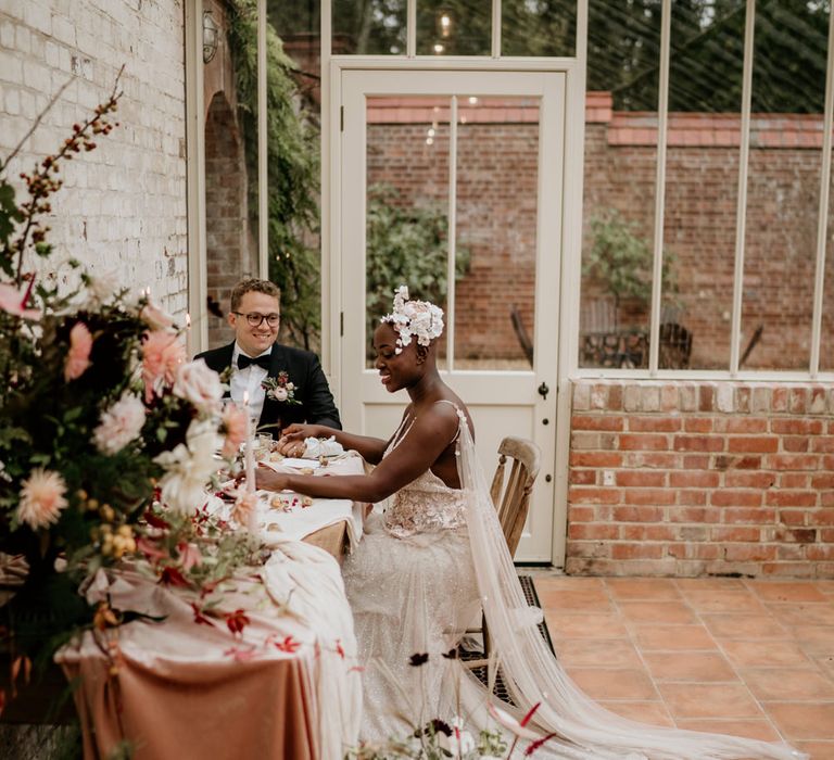 Bride with alopecia sitting at her intimate wedding breakfast in Our Beautiful Glasshouse in Sussex in a blush Julita Bridal London with watteau train 