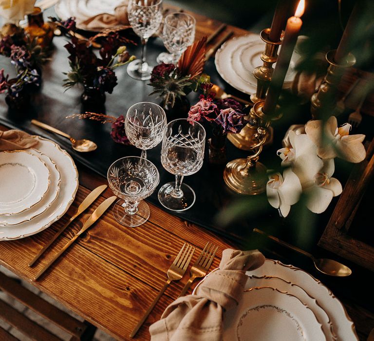 Place setting with ornate tableware, gold cutlery cut glassware and moody dark wedding flowers 