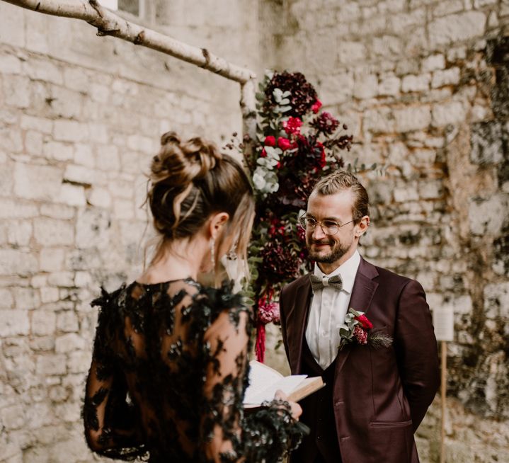 Groom in a burgundy three-piece wedding suit with bow tie smiling as his bride reads her vows to him