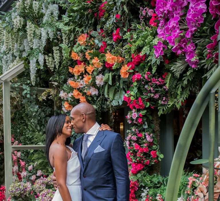 Portrait of a bride in a fitted Roland Mouret dress embracing her husband in a navy suit outside a flower shop in Chelsea 