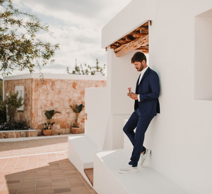 Stylish groom in a navy suit and white trainers leaning agains the Spanish villa wall 