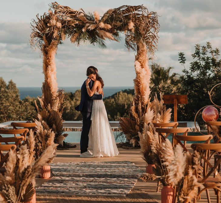 Boho bride and groom standing at the poolside altar under a dried flower arch 