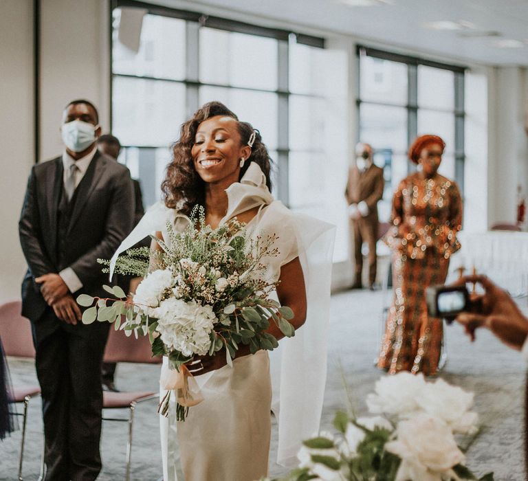 Smiling bride in customised white Bec + Bridge wedding dress with shoulder bows, white and green bouquet and curled wedding hair reaches the altar at Bridge Community Church wedding