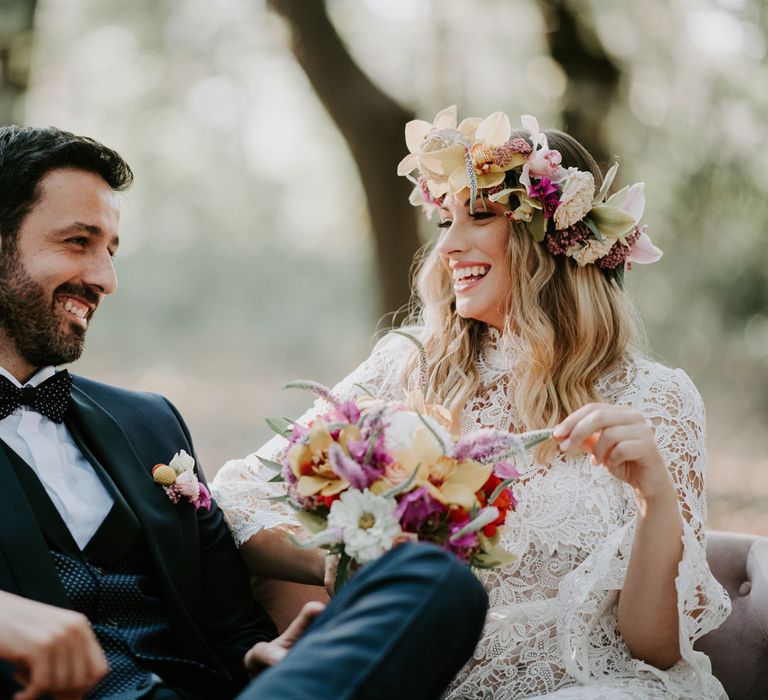 Bride in long lace boho wedding dress and flower crown sits on outside day bed swing in forest with groom in navy suit and bow tie at enchanted forest wedding in Italy