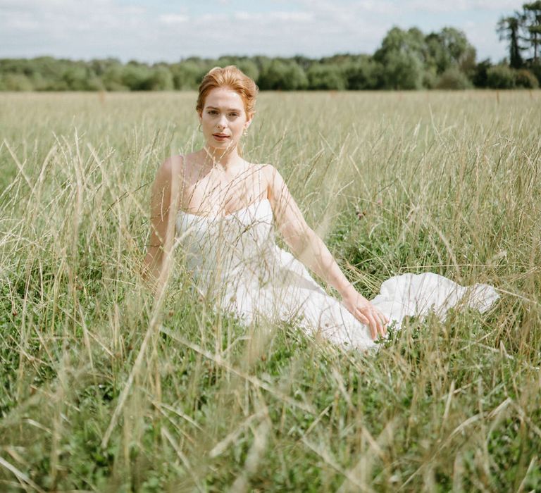 Bride sitting in the fields at Berwick Lodge wearing a spaghetti strap wedding dress, relaxed low pony tail and gold earrings